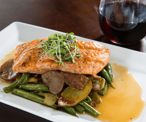 A plate of salmon over veggies. A glass of red wine sits on the table next to it.