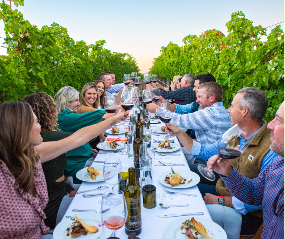 A group of people sitting in an Idaho winery vineyard are toasting with Idaho wine and food in front of them.